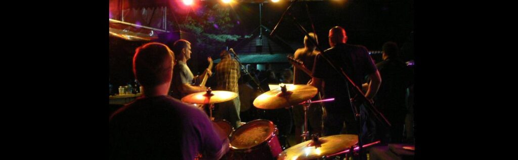 Musicians performing on stage with drum set in foreground and colorful stage lights.