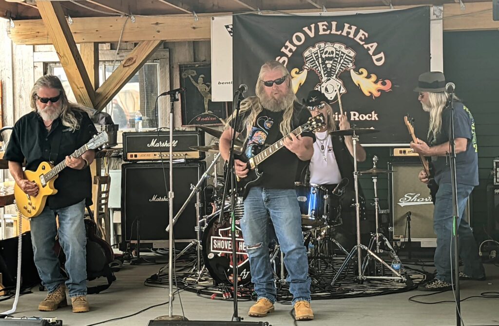 Musician playing bass guitar on stage with a Shovelhead banner behind.